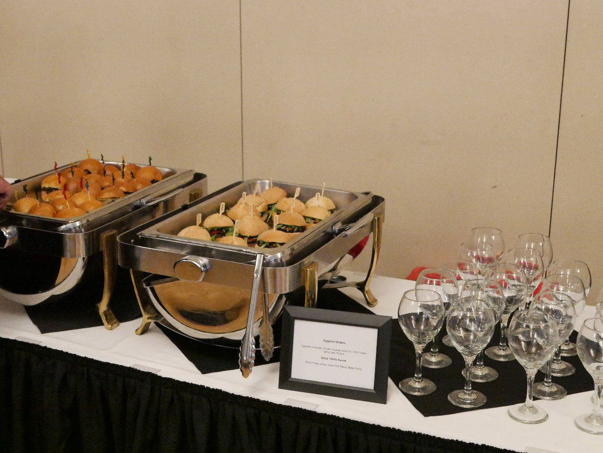 Two warming trays full of mini burgers sit next to a collection of wine glasses on a table with white linens.